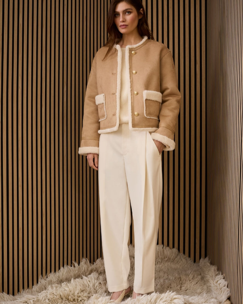 Model wearing camel faux leather jacket layered over knit and off white trousers